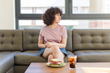 Genç, güzel bir Arap kadın evdeki kanepede sandviç yiyor.