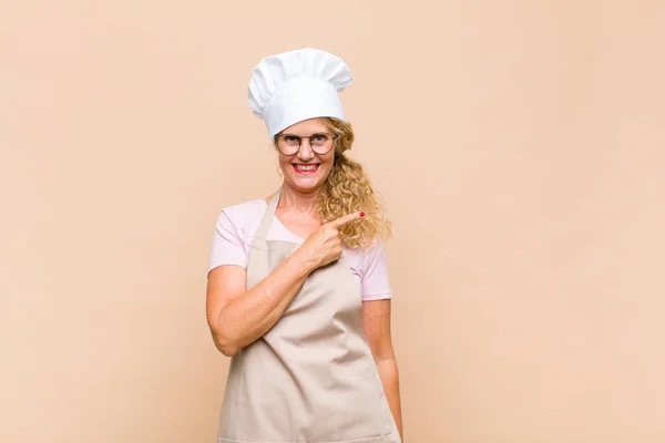 middle age woman baker smiling cheerfully, feeling happy and pointing to the side and upwards, showing object in copy space