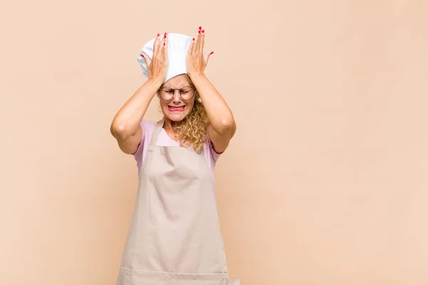 middle age woman baker feeling stressed and anxious, depressed and frustrated with a headache, raising both hands to head