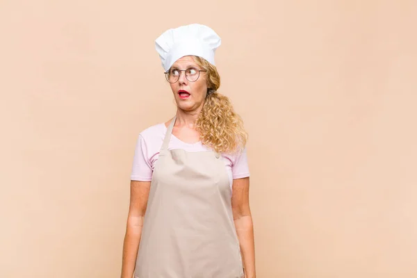middle age woman baker feeling shocked, happy, amazed and surprised, looking to the side with open mouth