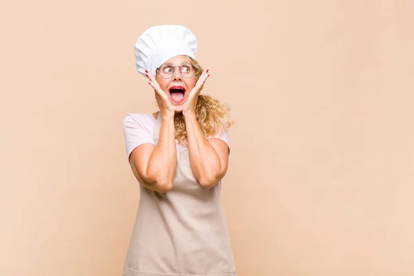 middle age woman baker feeling happy, excited and surprised, looking to the side with both hands on face