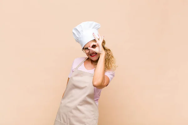 middle age woman baker smiling happily with funny face, joking and looking through peephole, spying on secrets