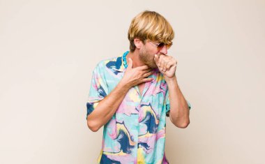 Sarışın, beyaz bir adam boğazı ağrıyormuş ve grip belirtileri gösteriyormuş. Ağzı kapalı öksürüyormuş.