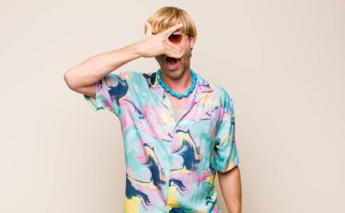 Sarışın, beyaz bir adam şok olmuş, korkmuş ya da korkmuş görünüyor. Yüzü elle örtülmüş ve parmaklarının arasından gizlice bakıyor.