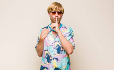 Sarışın, beyaz bir adam ciddi görünüyor ve parmaklarını dudaklarına bastırıp sessizlik ya da sessizlik talep ediyor, sır saklıyor.