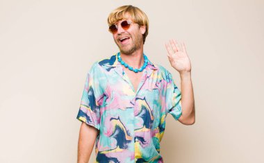 Sarışın, beyaz bir adam mutlu ve neşeli bir şekilde gülümsüyor, el sallıyor, seni selamlıyor ya da veda ediyor.