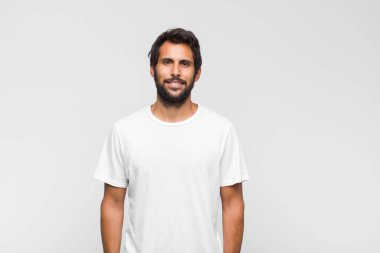 Genç yakışıklı Latin adam beyaz duvara poz veriyor ve kameraya bakıyor.