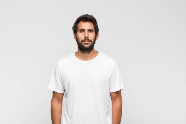 Genç yakışıklı Latin adam beyaz duvara poz veriyor ve kameraya bakıyor.