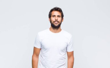 Genç yakışıklı Latin adam beyaz duvara poz veriyor ve kameraya bakıyor.