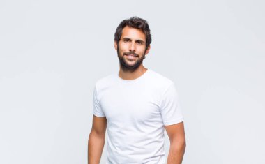 Genç yakışıklı Latin adam beyaz duvara poz veriyor ve kameraya bakıyor.