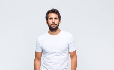 Genç yakışıklı Latin adam beyaz duvara poz veriyor ve kameraya bakıyor.