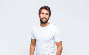 Genç yakışıklı Latin adam beyaz duvara poz veriyor ve kameraya bakıyor.