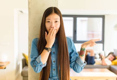 Genç, güzel Asyalı kadın kendini mutlu, şok olmuş ve şaşırmış hissediyor. Ağzını eliyle kapatıyor ve yanal fotokopi alanını işaret ediyor.