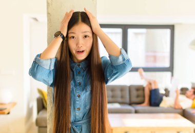 Genç, güzel Asyalı bir kadın heyecanlı ve şaşırmış görünüyor, ağzı açık, iki eli de başında, kendini şanslı bir kazanan gibi hissediyor.