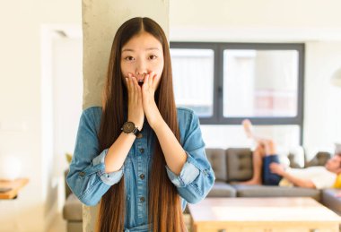 Genç, güzel Asyalı kadın şok olmuş ve korkmuş hissediyor, ağzı açık ve elleri yanaklarında korkmuş görünüyor.