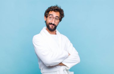 Banyo bornozu giymiş genç sakallı adam kameraya çapraz kollu gülümsüyor ve mutlu, kendinden emin, tatmin olmuş bir ifade çiziyor.