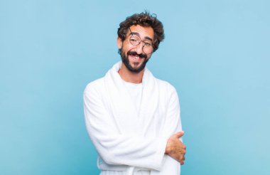 Banyo bornozu giymiş genç sakallı bir adam utangaç ve neşeli bir şekilde gülüyor, arkadaş canlısı ve olumlu ama kendine güveni olmayan bir tavırla