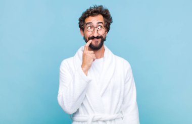 Genç sakallı adam banyo cübbesi giymiş mutlu bir şekilde gülümsüyor ve hayal kuruyor ya da kuşkulanıyor, yan tarafa bakıyor.