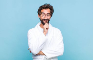 Banyo bornozu giymiş genç sakallı adam mutlu görünüyor ve eli çenesinde gülümsüyor, merak ediyor ya da soru soruyor, seçenekleri karşılaştırıyor.