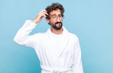 Banyo bornozu giymiş genç sakallı adam kafası karışmış, başını kaşıyor ve yan tarafa bakıyor.