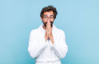 Banyo bornozu giyen genç sakallı adam mutlu, neşeli, şanslı ve iki eliyle ağzını kapatırken şaşırmış görünüyor.