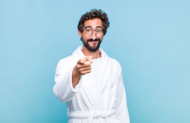 Banyo bornozu giymiş genç sakallı adam kameraya bakıyor. Tatmin olmuş, kendine güvenen, dost canlısı bir gülümsemeyle seni seçiyor.