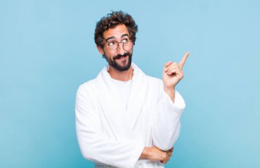 Genç sakallı bir adam banyo cübbesi giyiyor mutlu bir şekilde gülümsüyor ve yan bakıyor, merak ediyor, düşünüyor ya da bir fikri var.