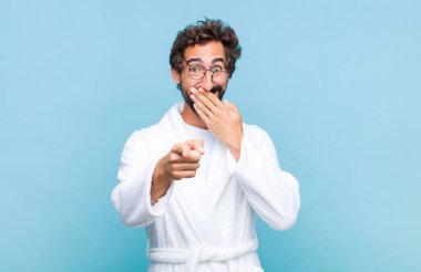 Banyo bornozu giymiş genç sakallı adam sana gülüyor, kamerayı işaret ediyor ve seninle dalga geçiyor ya da alay ediyor.