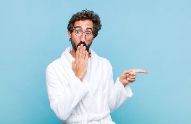 Banyo bornozu giyen genç sakallı adam mutlu, şok olmuş ve şaşırmış hissediyor. Ağzını eliyle kapatıyor ve yanal fotokopi alanını işaret ediyor.