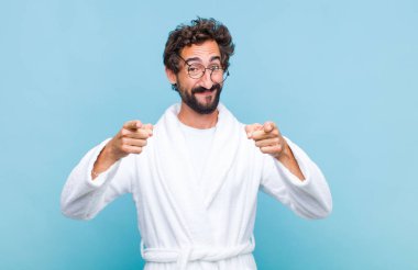 Genç sakallı adam banyo cübbesi giyiyor mutlu ve kendinden emin, kameraya iki eliyle bakıyor ve gülüyor, seni seçiyor.