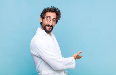 Genç sakallı bir adam banyo cübbesi giyiyor mutlu ve neşeli hissediyor, gülümsüyor ve seni selamlıyor, dostça bir jestle içeri davet ediyor.