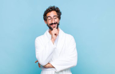 Banyo bornozu giymiş genç sakallı bir adam mutlu, kendinden emin bir ifadeyle gülümsüyor, çenesi üzerinde, merak ediyor ve yan tarafa bakıyor.