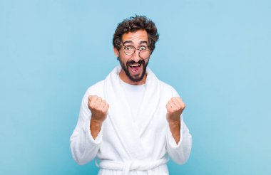 Banyo bornozu giyen genç sakallı adam şok olmuş, heyecanlı ve mutlu hissediyor, gülüyor ve başarıyı kutluyor.!