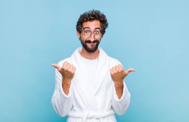 Banyo bornozu giymiş genç sakallı adam neşe içinde gülümsüyor ve mutlu görünüyor, tasasız ve pozitif hissediyor.