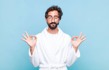 Banyo bornozu giymiş genç sakallı adam konsantre olmuş ve meditasyon yapıyor, tatmin olmuş ve rahatlamış hissediyor, düşünüyor ya da bir seçim yapıyor.