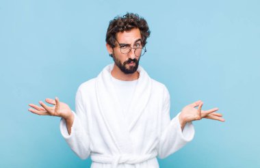 Banyo bornozu giyen genç sakallı adam şaşkın, kafası karışık ve stresli görünüyor, farklı seçenekler arasında merak ediyor, kararsız hissediyor.