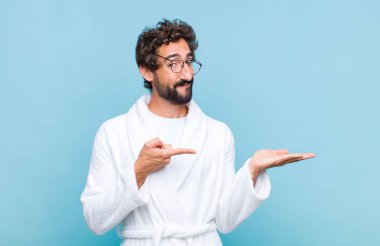 Genç sakallı adam banyo cübbesi giyiyor gülümsüyor, mutlu, kaygısız ve tatmin olmuş hissediyor, fotokopi alanındaki konsepte veya fikre işaret ediyor.