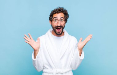 Banyo bornozu giyen genç sakallı adam mutlu, heyecanlı, şaşırmış ya da şok olmuş hissediyor, gülümsüyor ve inanılmaz bir şeye hayret ediyor.