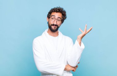 Banyo bornozu giymiş genç sakallı adam gururla ve gururla gülümsüyor, mutlu ve tatmin olmuş hissediyor ve fotokopi alanında bir konsept gösteriyor.