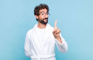 Banyo bornozu giymiş genç sakallı adam gururla gülümsüyor ve gururla bir numaralı pozunu veriyor, kendini lider gibi hissediyor.
