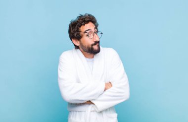 Genç sakallı bir adam banyo cübbesi giyiyor, omuz silkiyor, kafası karışık ve kararsız, kolları bağlı ve şaşkın görünüyor.