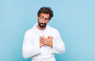 Banyo bornozu giymiş genç sakallı adam üzgün, yaralı ve kalbi kırık görünüyor, iki elini de kalbe yakın tutuyor, ağlıyor ve bunalımda.