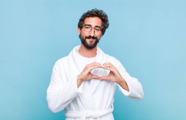 Genç sakallı bir adam banyo cübbesi giyiyor gülümsüyor ve mutlu, sevimli, romantik ve aşık, iki eliyle kalbi şekillendiriyor.
