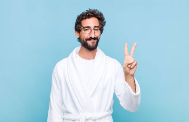Banyo bornozu giymiş genç sakallı bir adam gülümsüyor ve arkadaş canlısı görünüyor, iki ya da iki numarayı gösteriyor, eli önde, geri sayıyor.