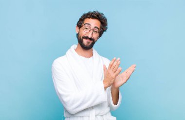 Banyo bornozu giymiş genç sakallı adam mutlu ve başarılı hissediyor, gülümsüyor ve alkışlıyor, alkışlarla tebrik ediyor.
