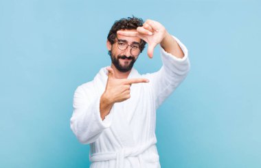 Banyo bornozu giyen genç sakallı adam mutlu, arkadaş canlısı ve pozitif hissediyor, gülümsüyor ve elleriyle bir portre veya fotoğraf çerçevesi yapıyor.