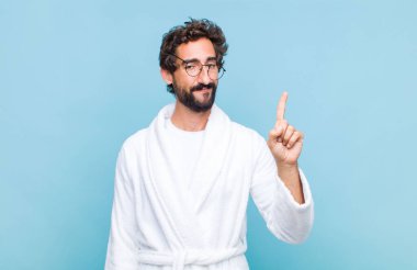 Genç sakallı bir adam banyo cübbesi giyiyor gülümsüyor ve arkadaş canlısı görünüyor, bir numarayı gösteriyor veya ilk eli önde, geri sayıyor.