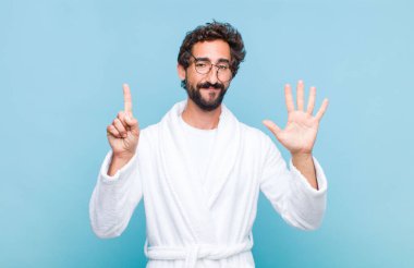 Banyo bornozu giymiş genç sakallı bir adam gülümsüyor ve arkadaş canlısı görünüyor, 6 ya da 6 numarayı gösteriyor, elleri önde, geriye doğru sayıyor.