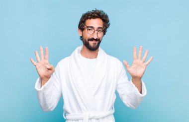 Banyo bornozu giymiş genç sakallı bir adam gülümsüyor ve arkadaş canlısı görünüyor, 9 ya da 9 numarayı gösteriyor, geriye doğru sayıyor.