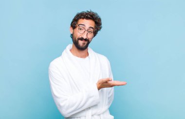 Genç sakallı bir adam banyo cübbesi giyip neşeli bir şekilde gülümsüyor, mutlu hissediyor ve fotokopi çekilen alanda avucuyla bir konsept gösteriyor.